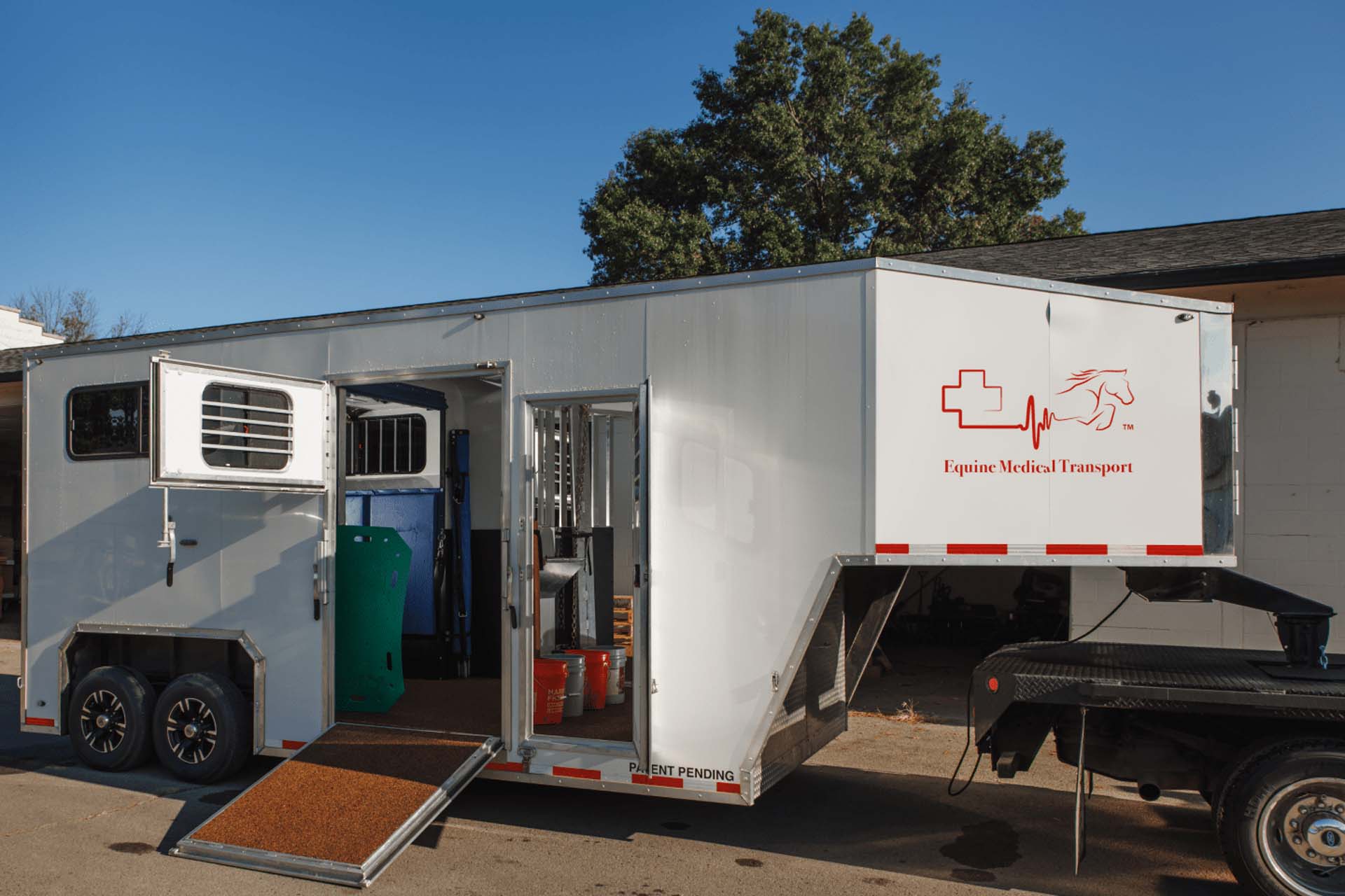 A picture of a State-of-the-art Equine Ambulance. It’s a white trailer hitched to the back of a black truck that has “Equine Medical Transport” printed on it.